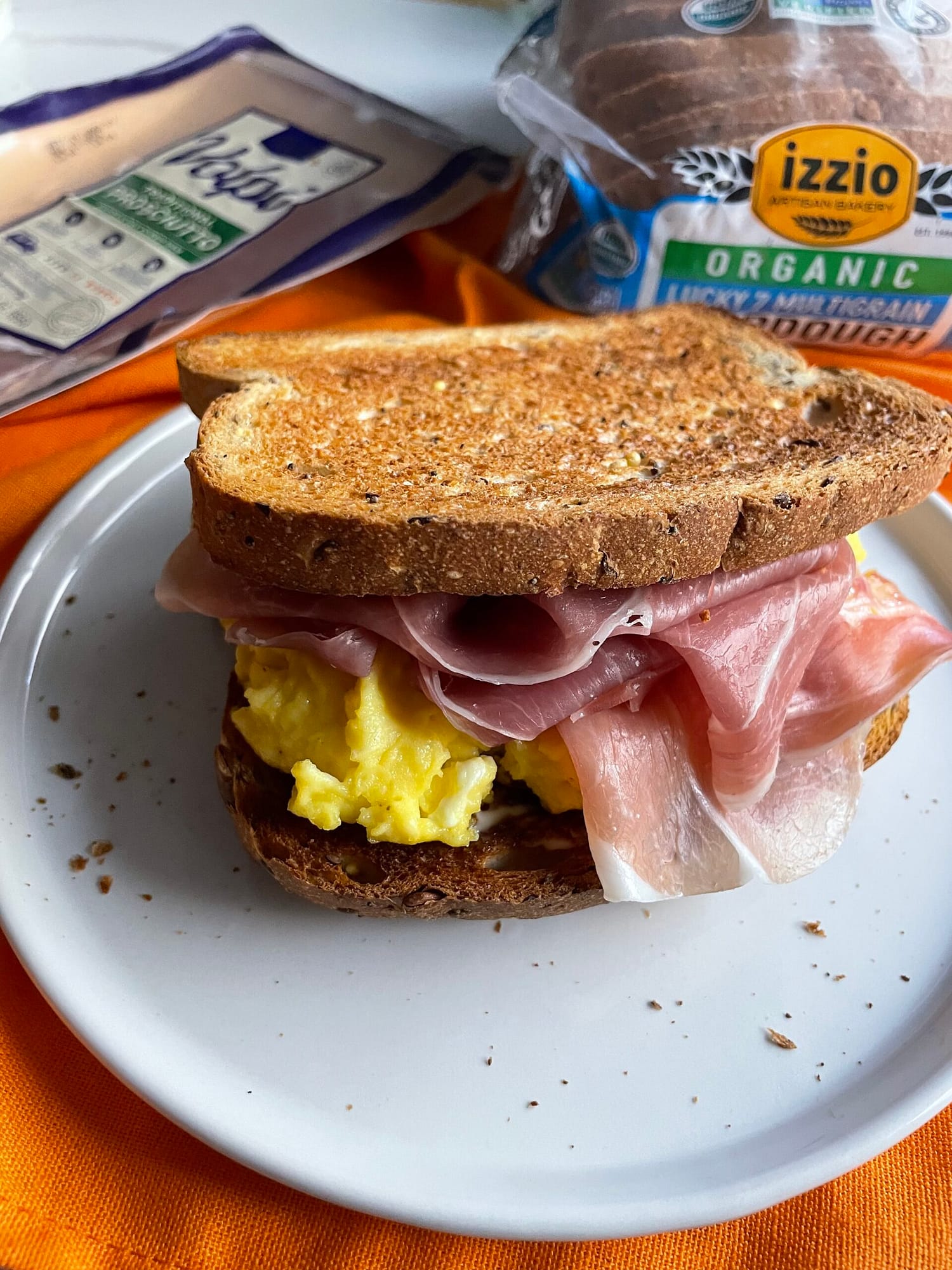 Sandwich on izzio Lucky Seven Multigrain Sourdough with Volpi brand prosciutto and scrambled eggs pictured on white plate