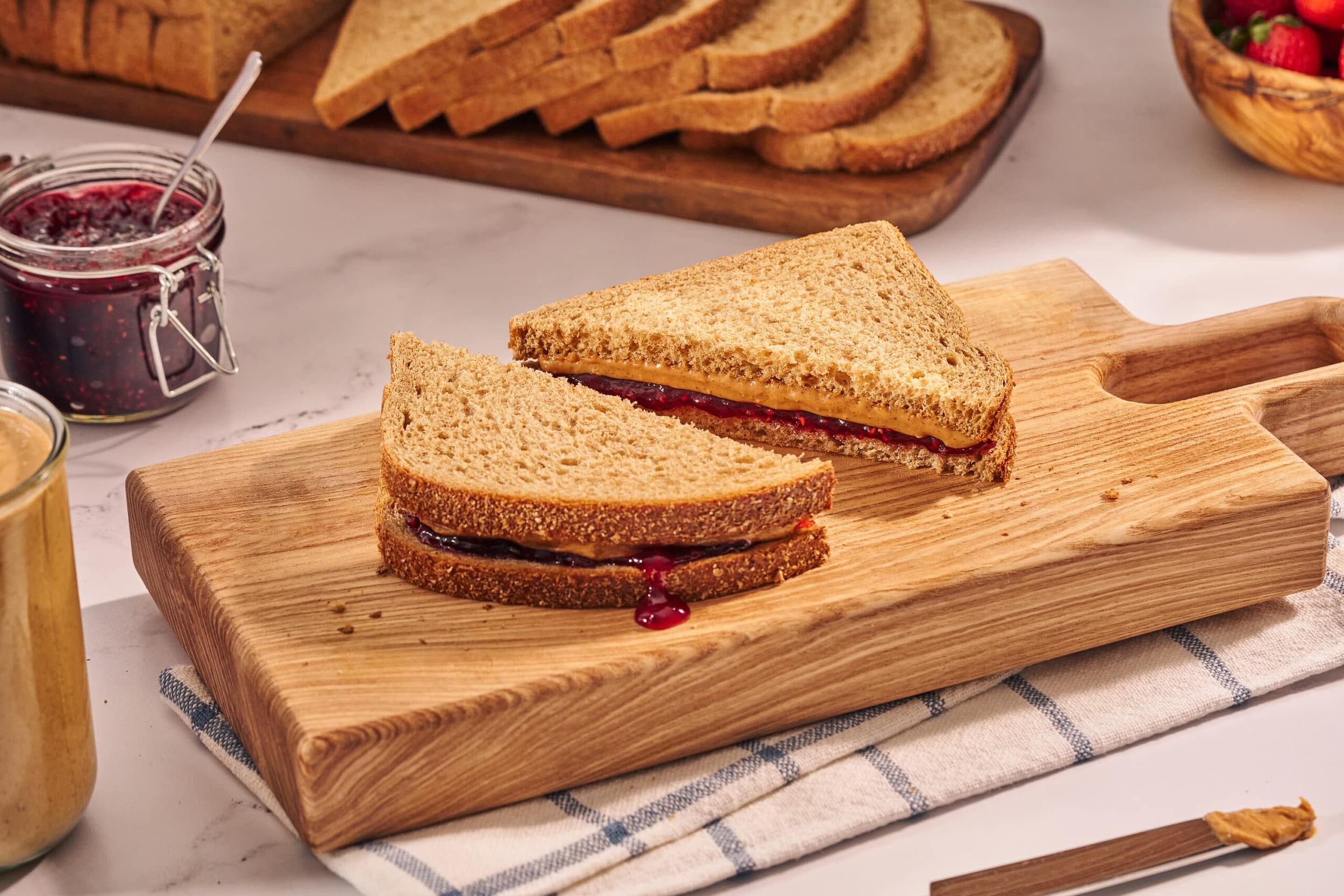 peanut butter and jelly on izzio organic whole wheat sandwich bread stacked on a cutting board, layered on a towel with peanut butter, jelly, and extra bread slices around