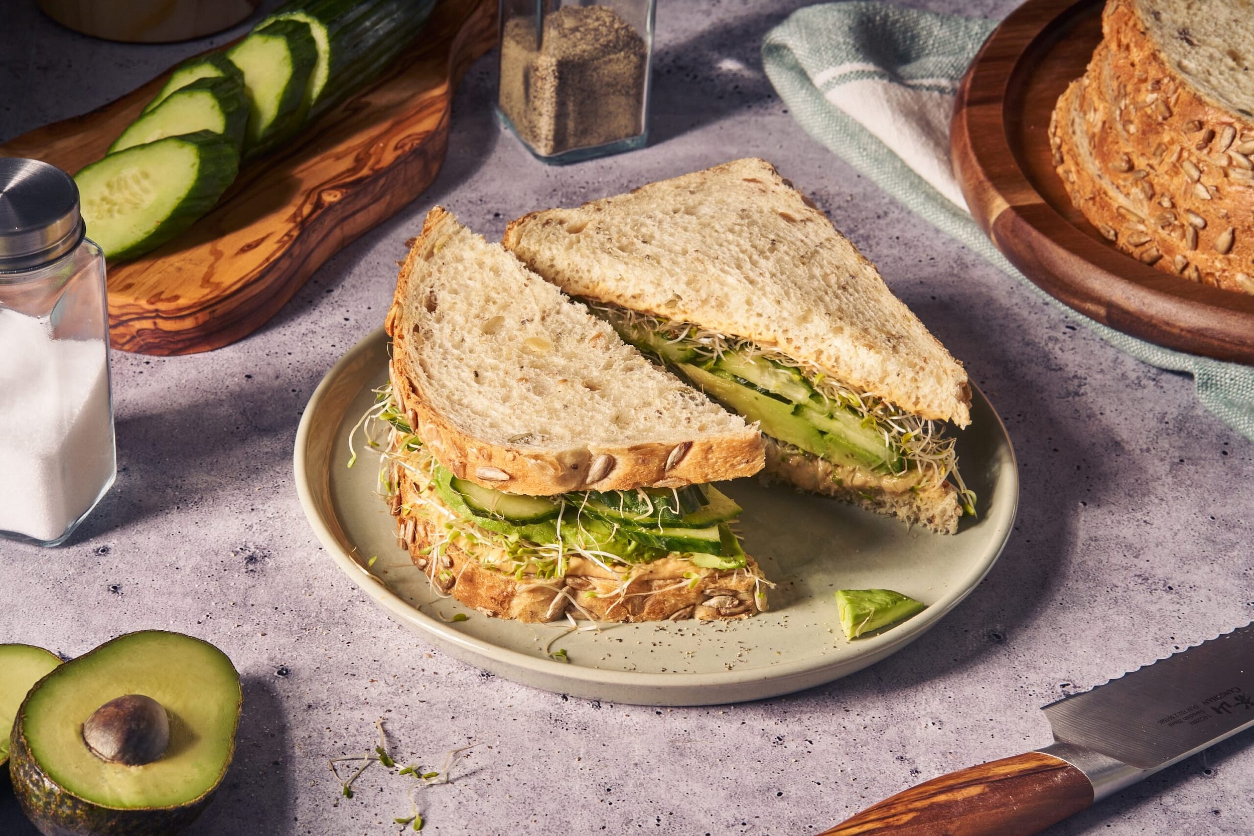 Avocado, sprouts, and greens Sandwich on a seedy, grain-dense sourdough bread placed on white plate with avocado, cucumber surrounding the plate